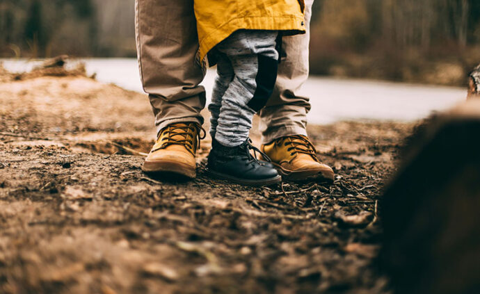 a young child stands between a parents legs on top of soil in a forest clearing