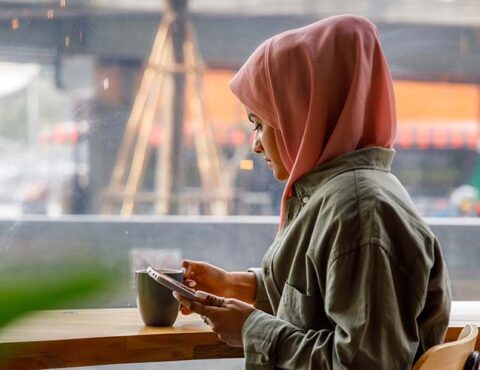 Arabic businesswoman using mobile phone while drinking coffee at office cafe. She is wearing headscarf sitting at coffee shop