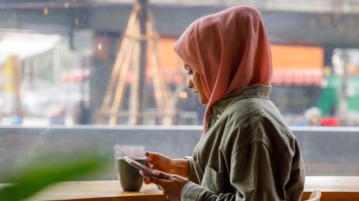 Arabic businesswoman using mobile phone while drinking coffee at office cafe. She is wearing headscarf sitting at coffee shop