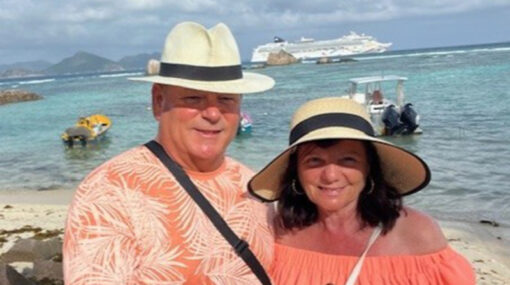 Photo of Lorraine Russell with her husband Craig on holiday stood on a beach with a cruise ship in the sea in the background